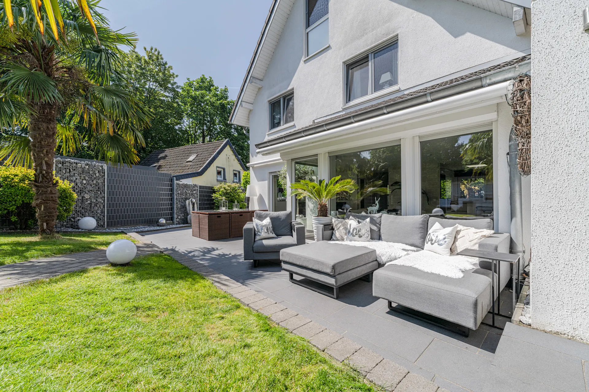 Moderne Terrasse mit grauem Sofa, Palmbaum und Blick auf weißen Wohnhausgarten.