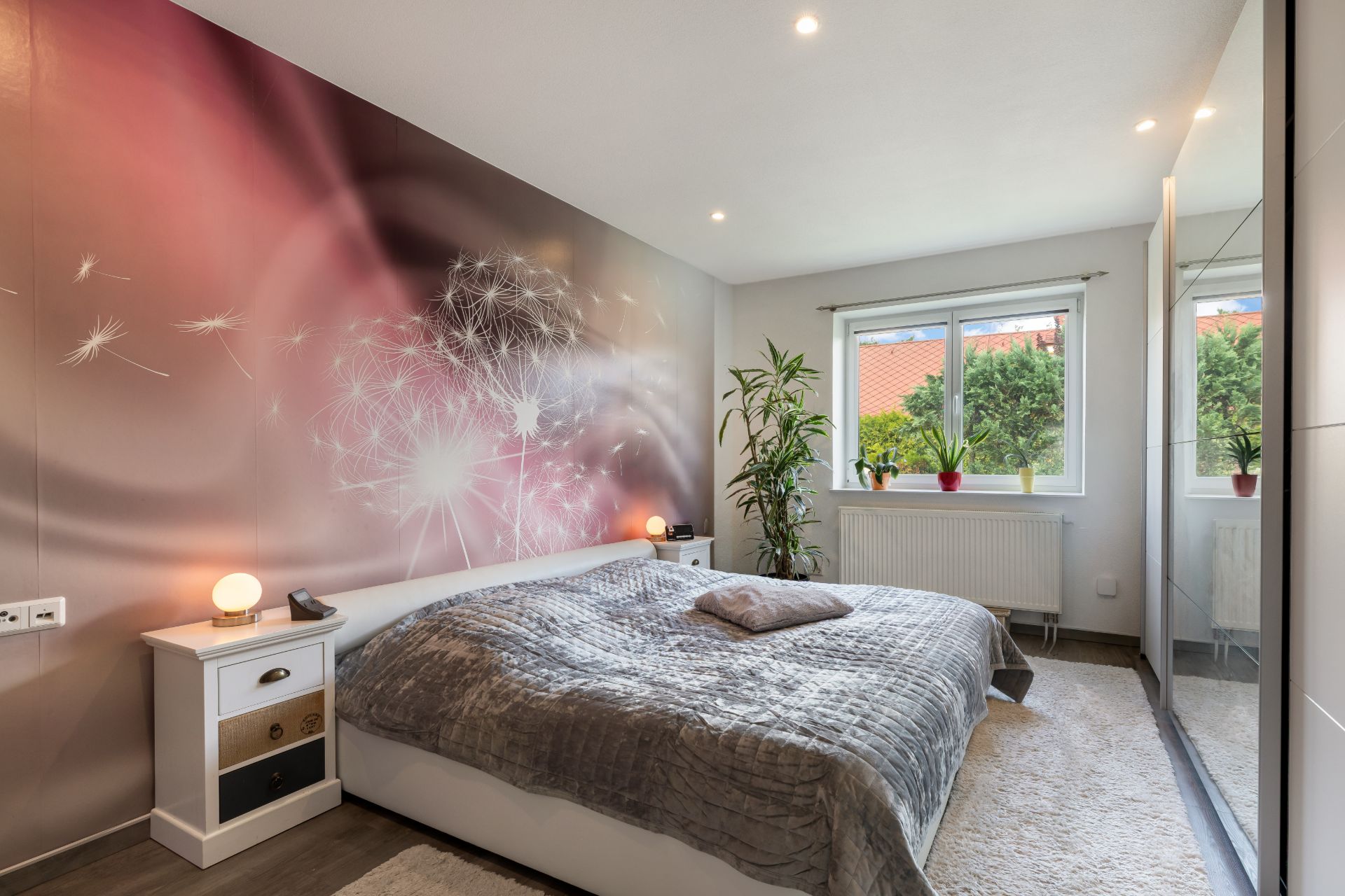 Modernes Schlafzimmer mit großem Bett, Pflanzen, Wandbild und Fenster mit Blick ins Grüne.