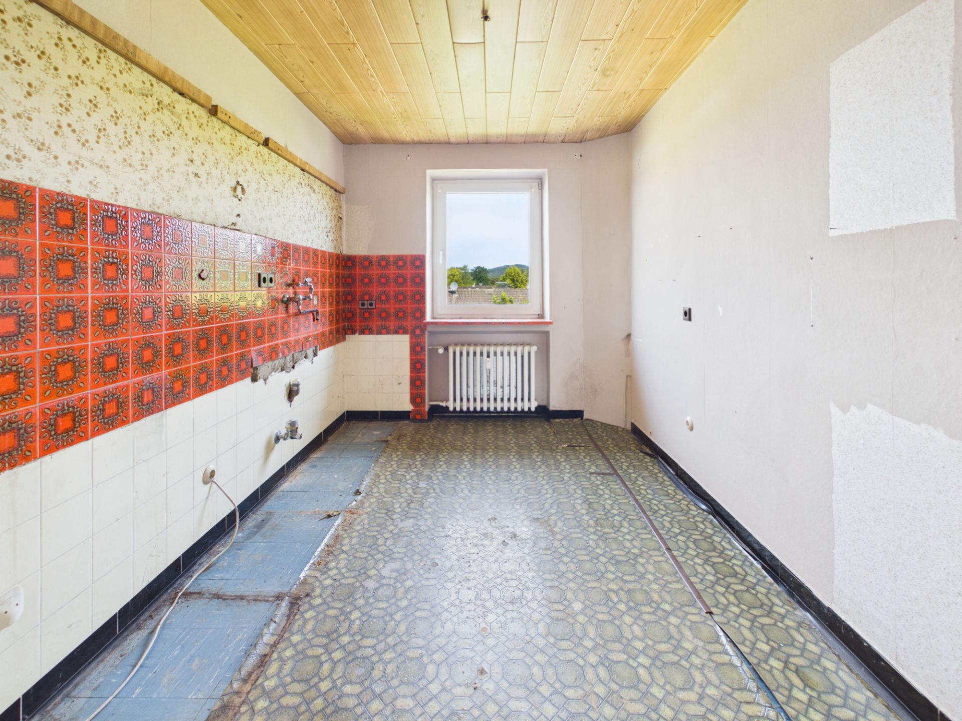 Leere Küche mit bunten Wandfliesen, Fenster und Heizkörper am Ende des Raums.