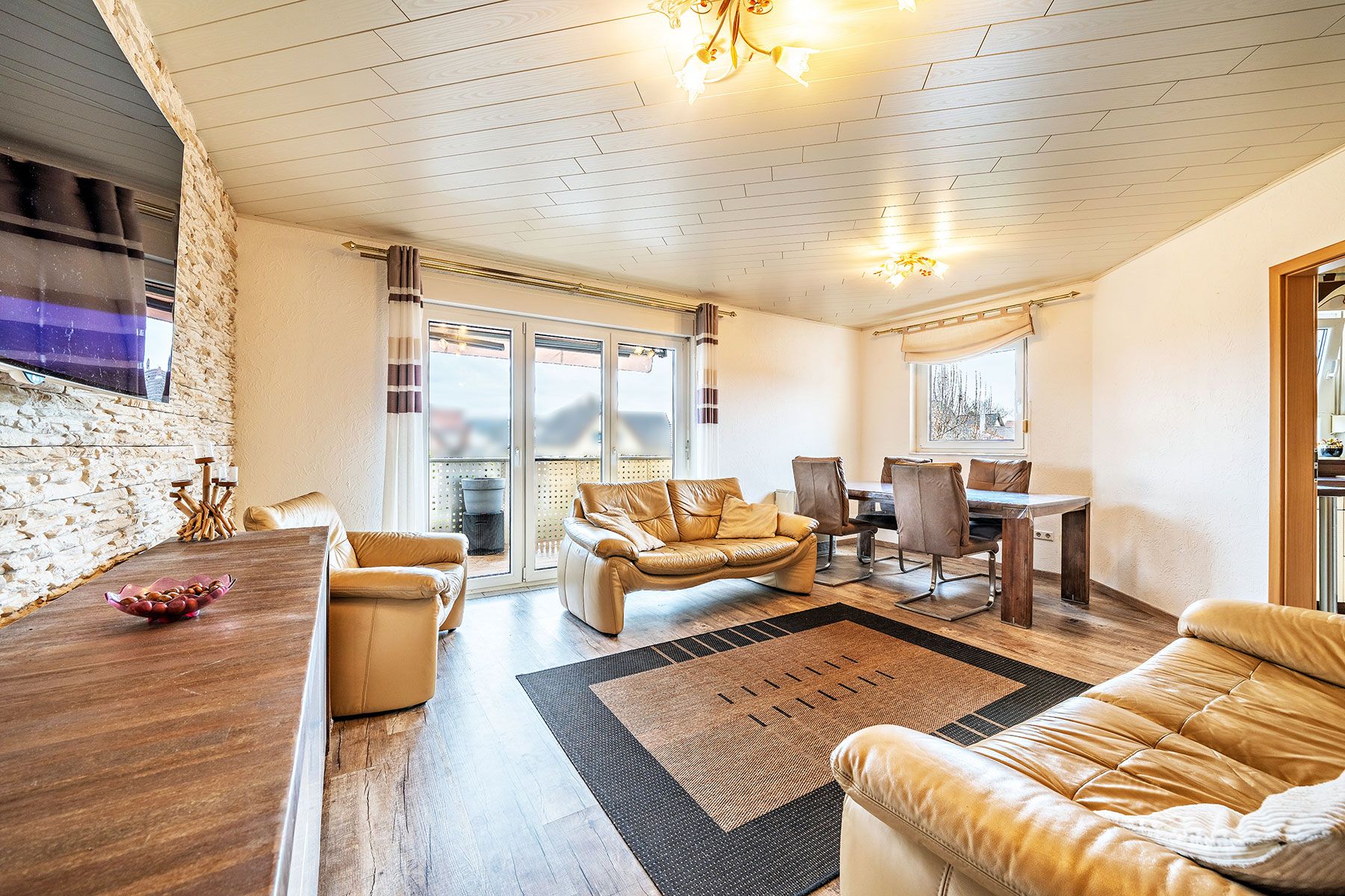 Helles Wohnzimmer mit Ledersofas, Esstisch, großem Teppich und Zugang zum Balkon.
