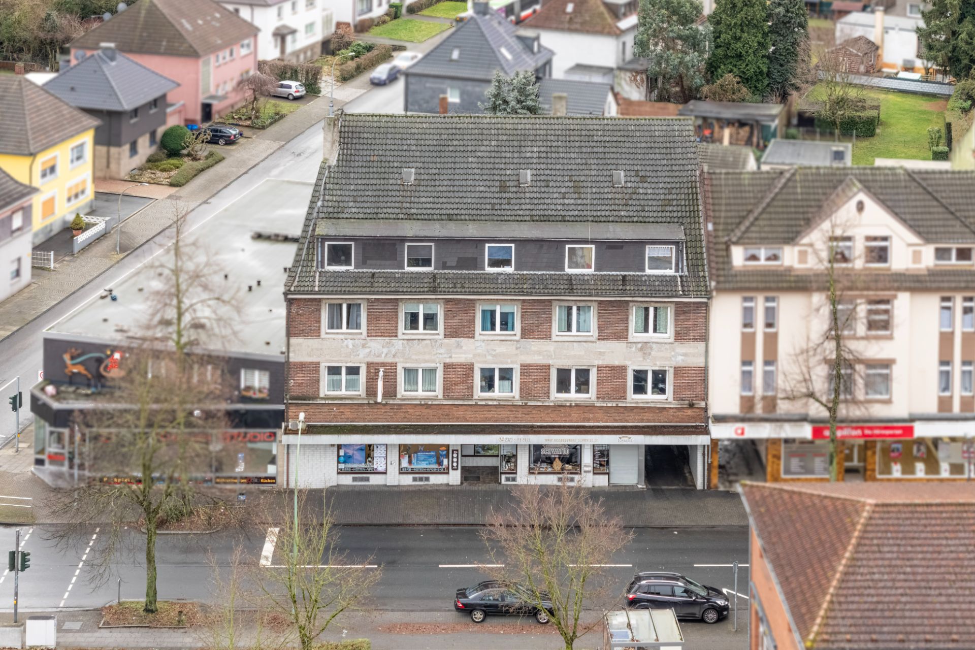 Mehrstöckiges Wohngebäude mit Geschäften im Erdgeschoss an einer Straße, Bäume und Autos davor.