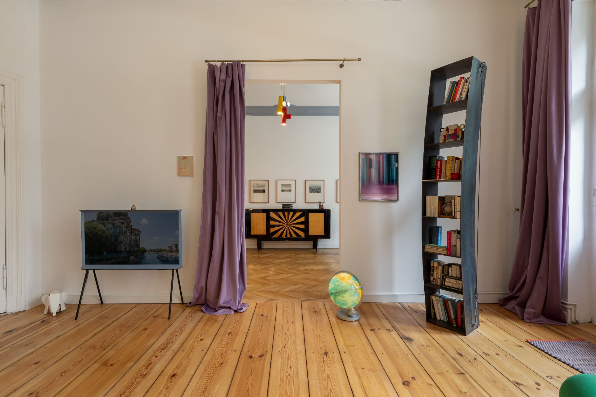 Wohnzimmer mit Holzfußboden, Bücherregal, Globus und lila Vorhängen.