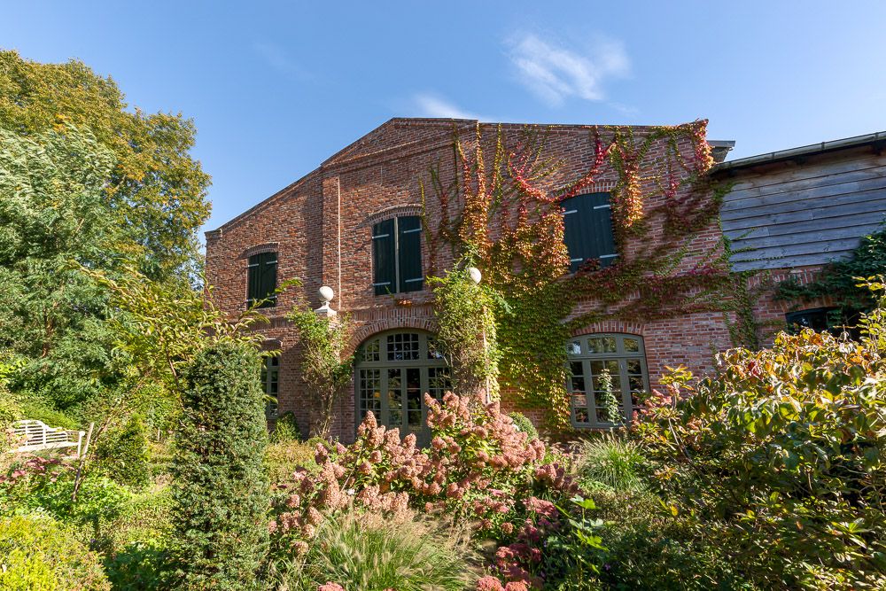 Backsteinhaus mit vielen Fenstern, bewachsene Fassade und üppigem Garten im Vordergrund.