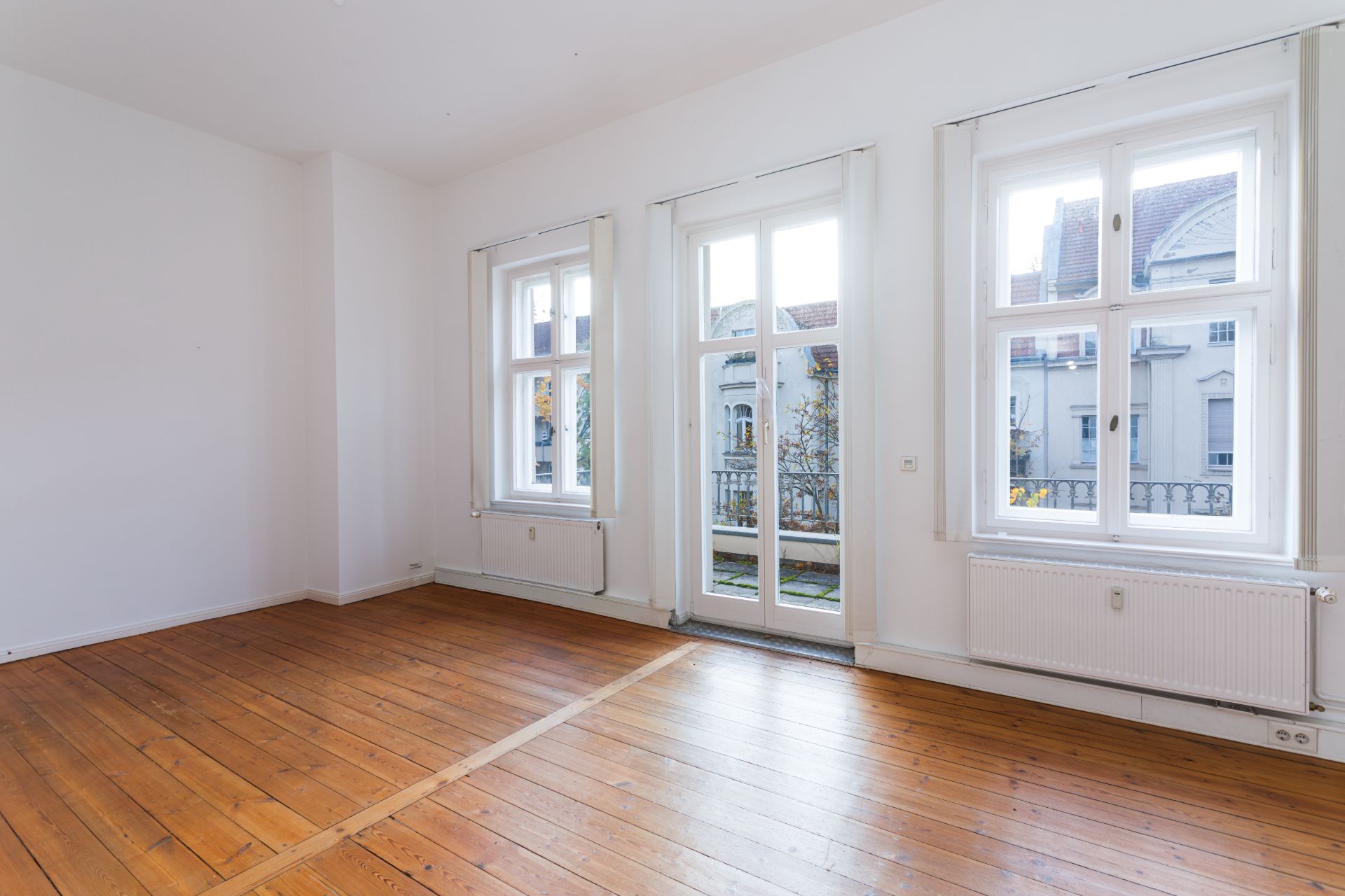 Heller leerer Raum mit Holzboden, drei Fenstern und einer Tür zum Balkon.