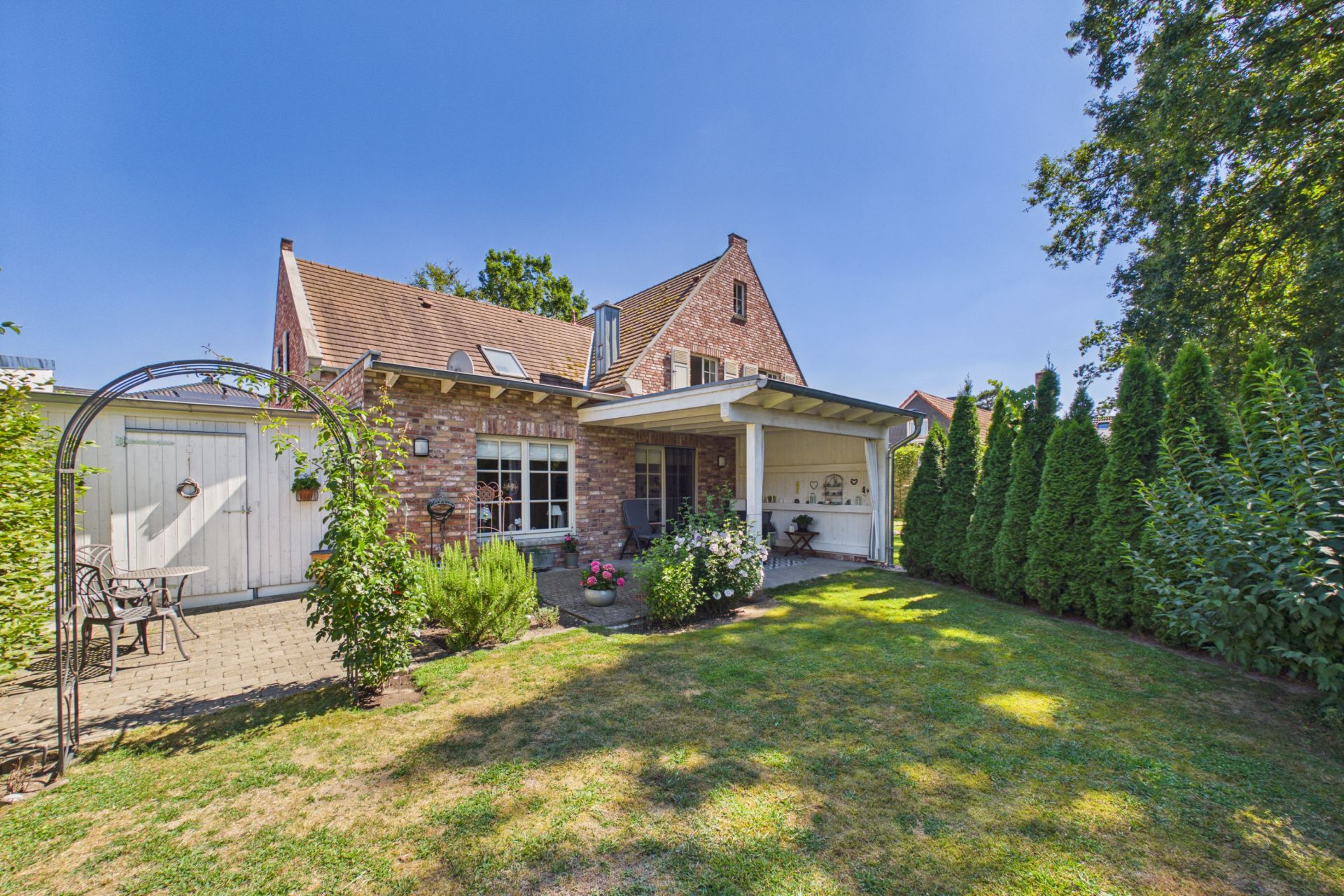 Backsteinwohnhaus mit überdachter Terrasse, Garten und Sitzbereich im Freien, umgeben von Bäumen.