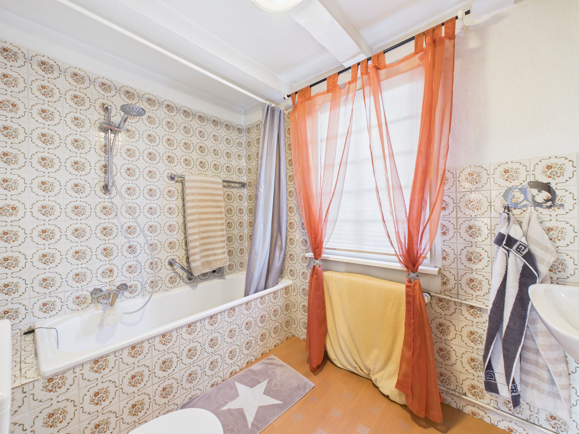 Badezimmer mit Badewanne, gemusterten Fliesen und orangefarbenem Vorhang am Fenster.
