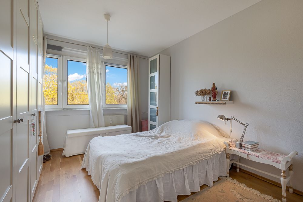 Helles Schlafzimmer mit großem Bett, Kleiderschrank und drei Fenstern, Holzboden und weißer Einrichtung.