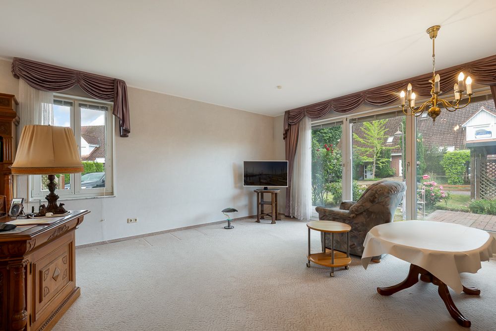 Helles Wohnzimmer mit Sessel, rundem Tisch, TV und Blick durch große Fenster in den Garten.