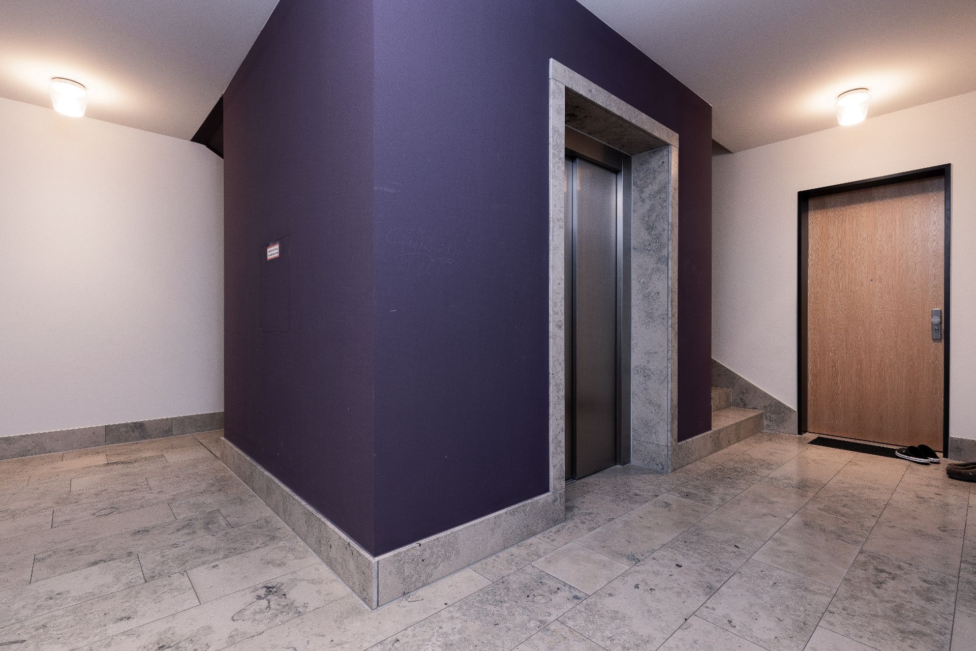 Flur mit Aufzug, lila Wand, Haustür und Treppe mit Fliesenboden.