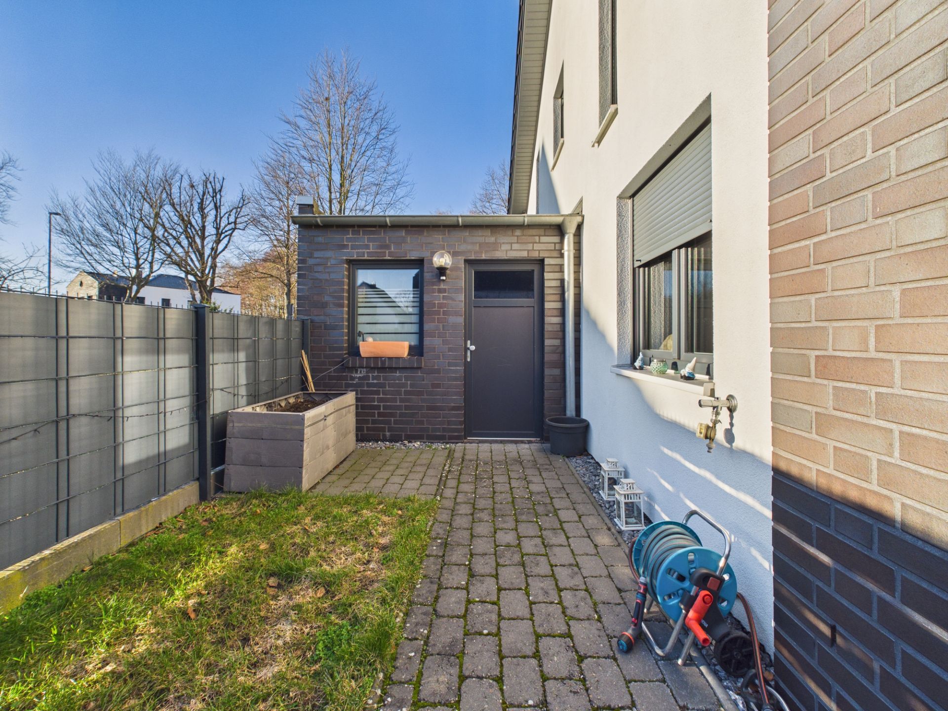Kleiner gepflasterter Hinterhof mit Rasen, Hochbeet und Gartenschlauch an einem Wohnhaus.