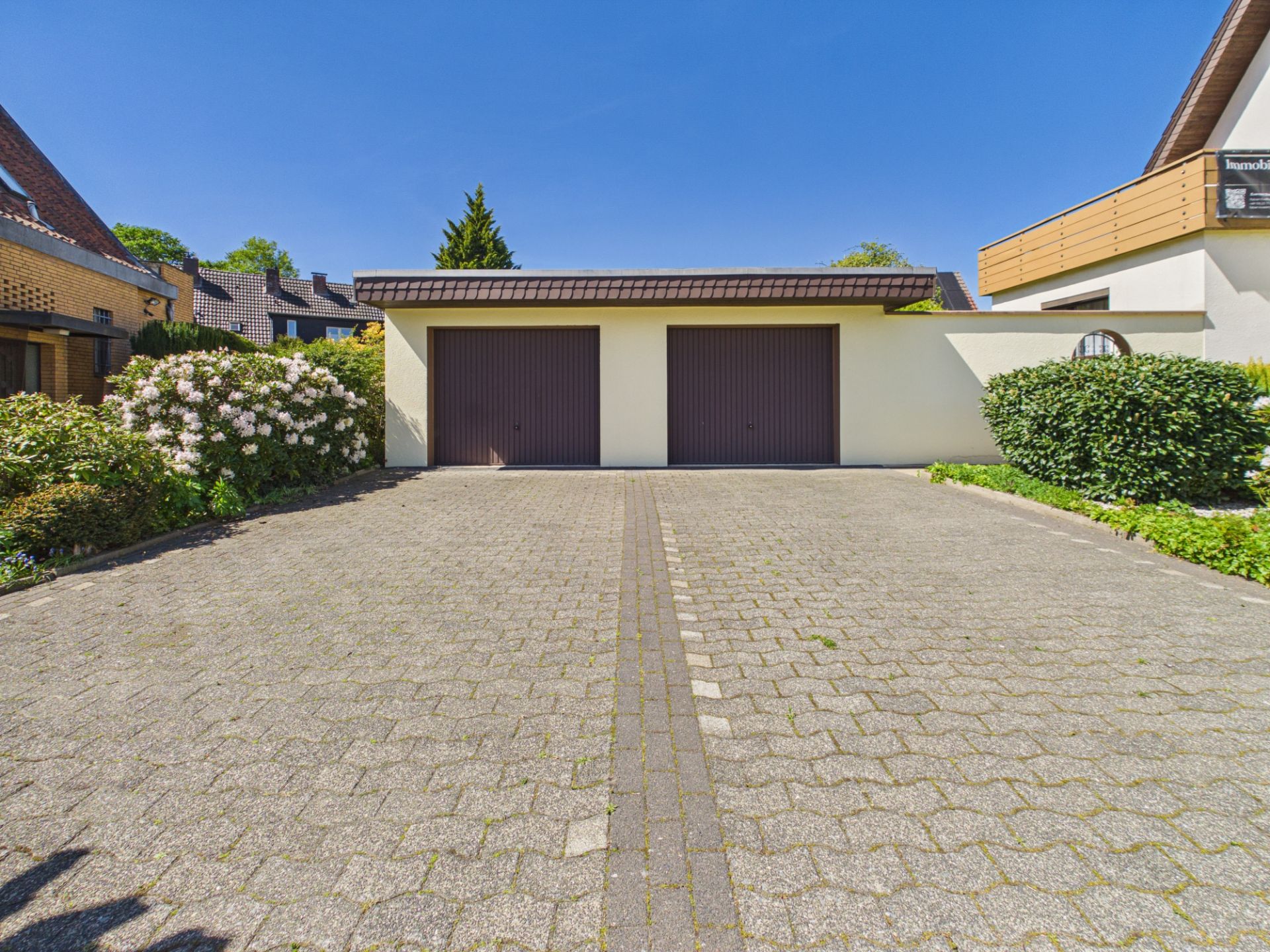 Doppelgarage mit zwei Toren und gepflasterter Einfahrt, umgeben von Büschen und Wohnhäusern.