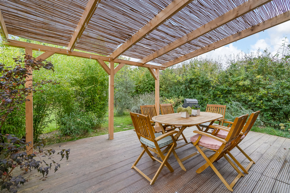 Überdachte Holzterrasse mit Tisch und sechs Stühlen im Garten, umgeben von grünen Pflanzen.