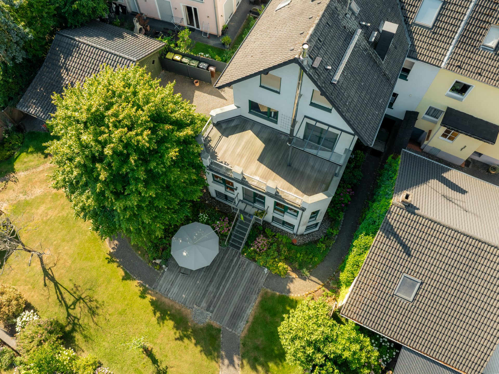 Luftaufnahme eines Wohnhauses mit Holzterrasse, Garten und großem Baum.