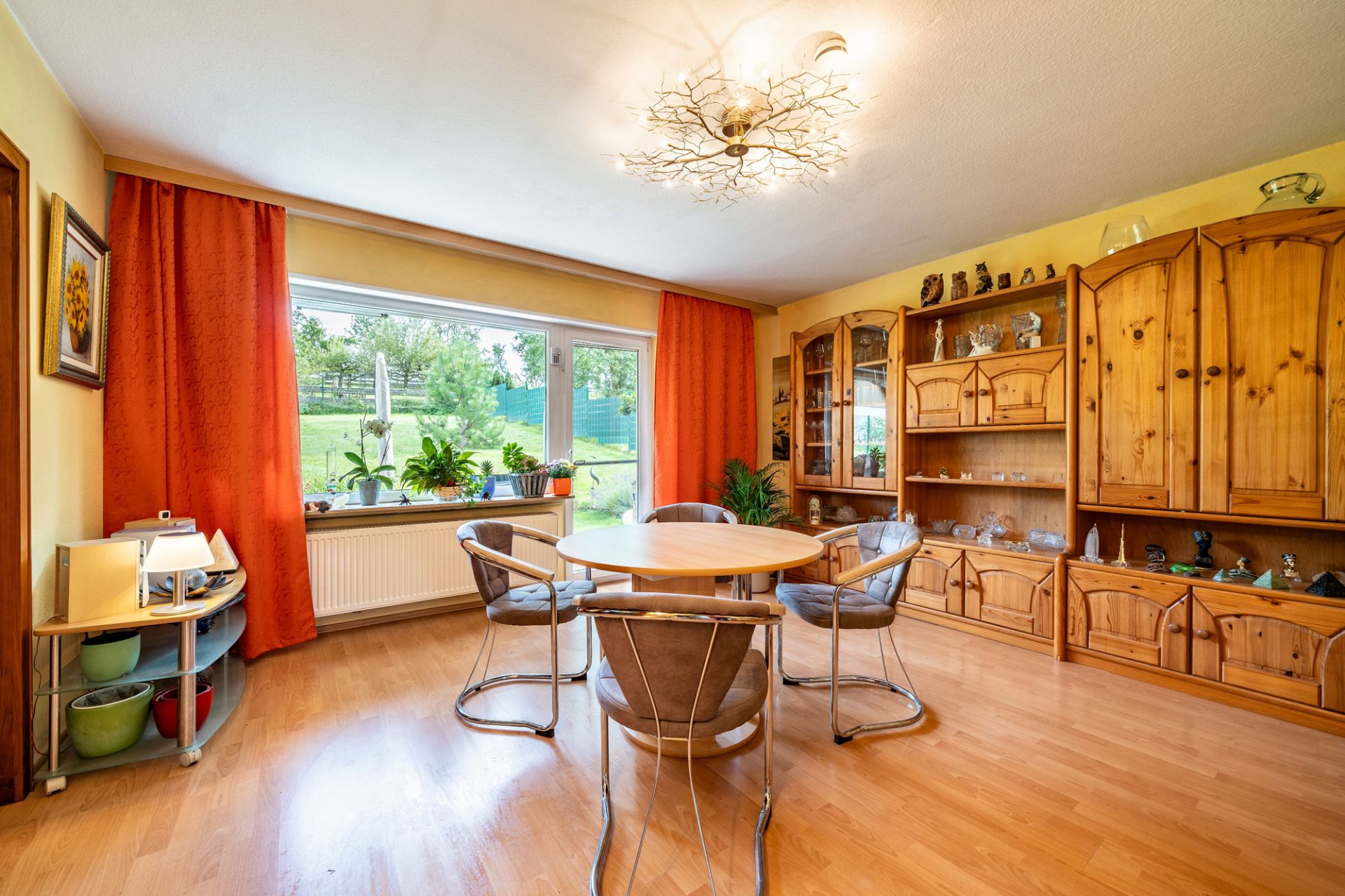 Helles Wohnzimmer mit rundem Tisch, vier Stühlen, Holzschränken und großem Fenster mit orangefarbenen Vorhängen.