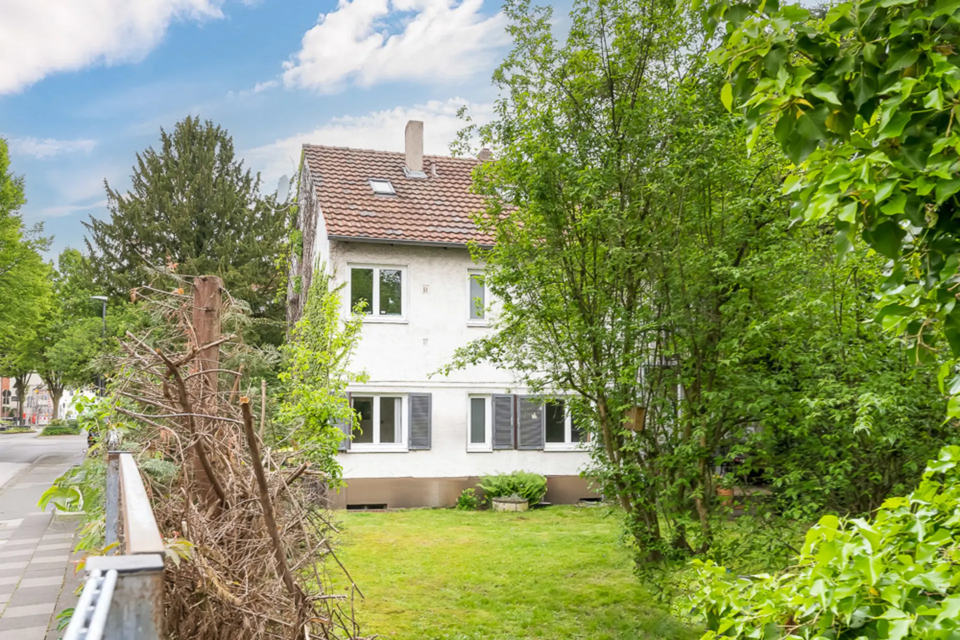 Weißes Wohnhaus mit rotem Dach, Garten und vielen grünen Bäumen im Vordergrund.
