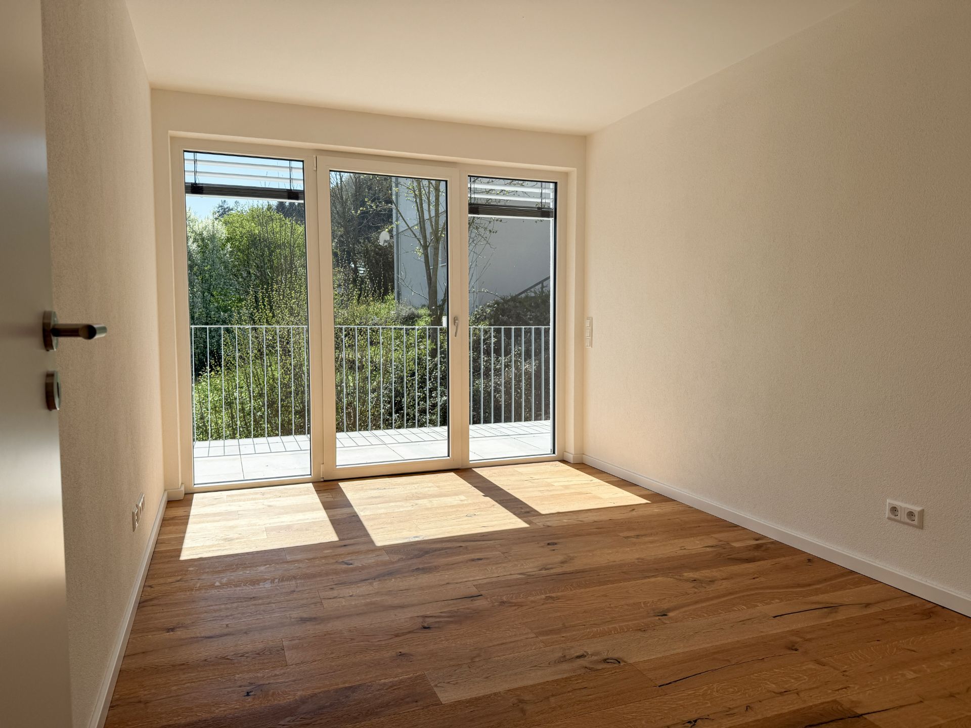 Heller leerer Raum mit Holzfußboden und großer Fensterfront zu einem Balkon.