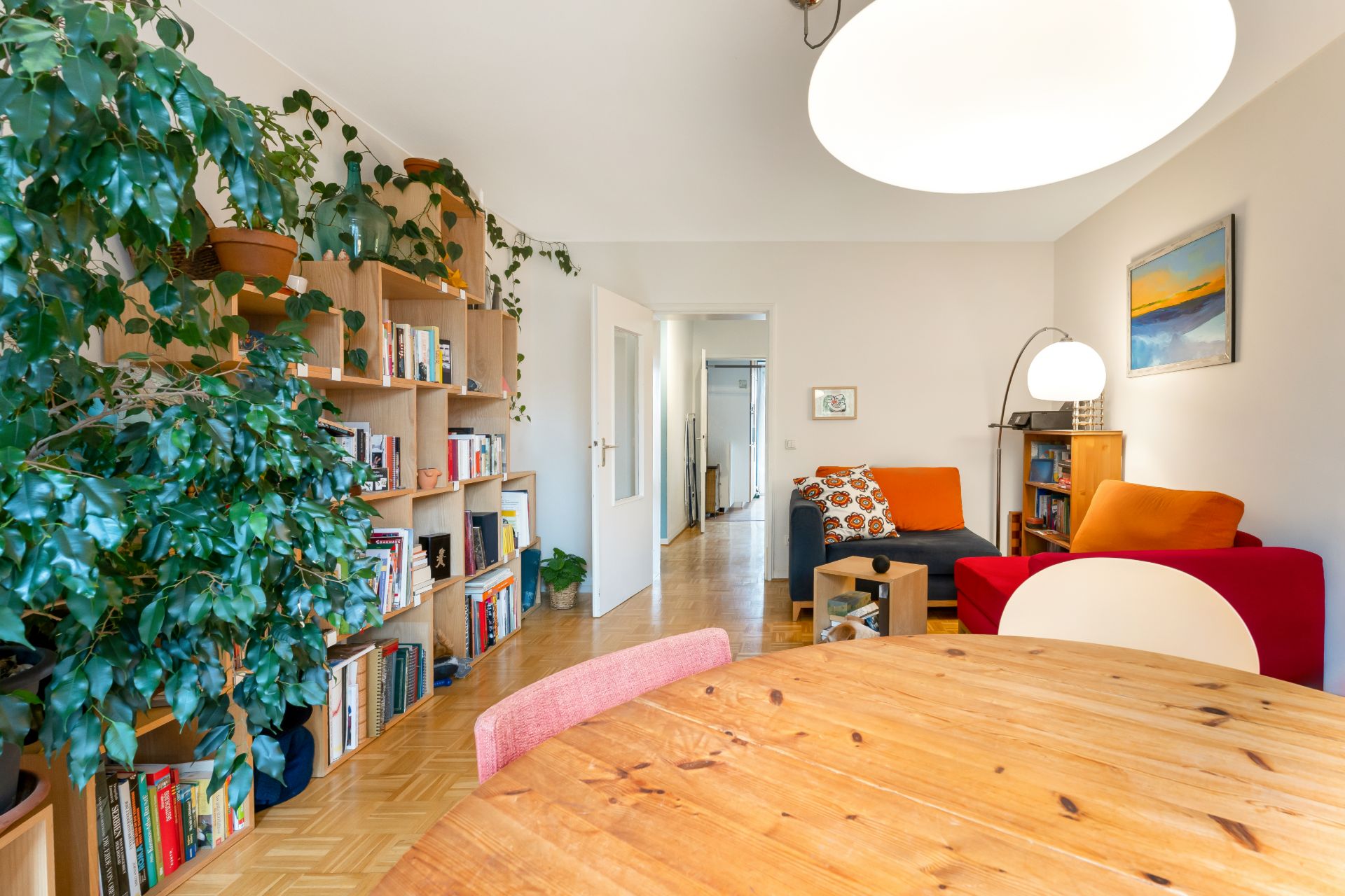 Helles Wohnzimmer mit Bücherregal, großen Pflanzen, Holztisch und rotem Sofa.