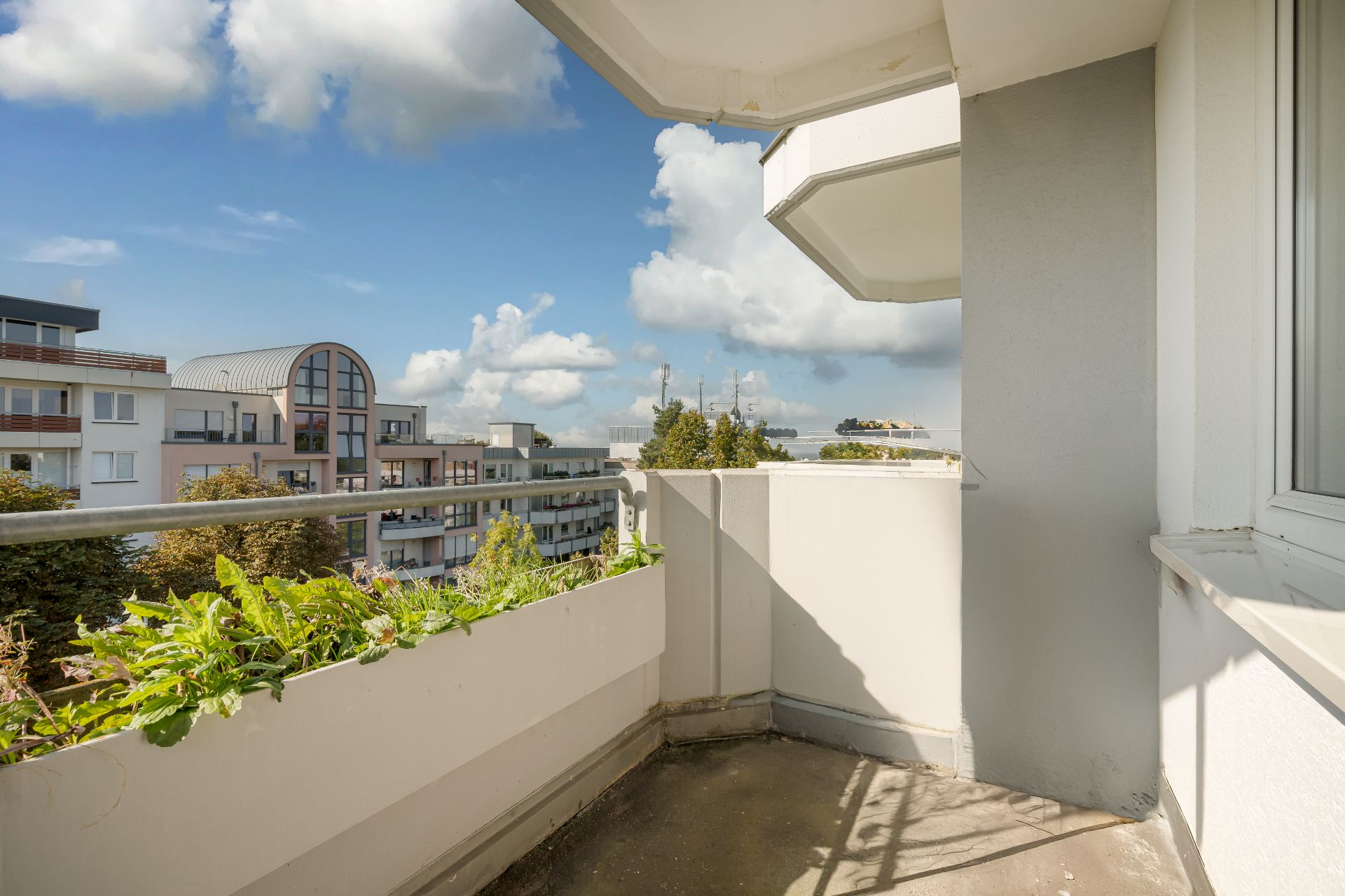 Balkon mit Pflanzenkasten und Blick auf moderne Wohngebäude unter blauem Himmel.
