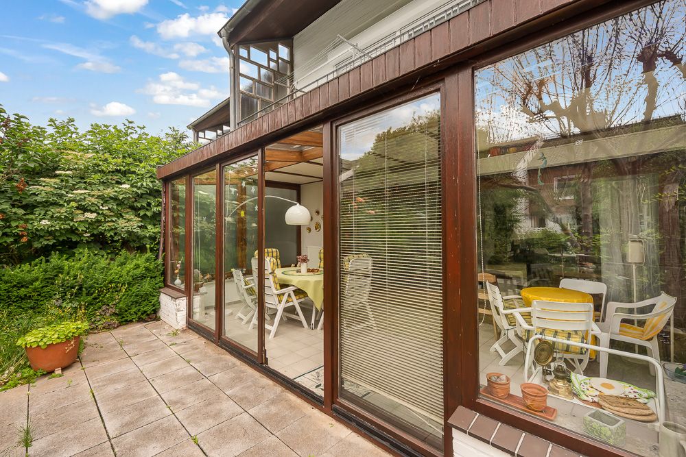 Wintergarten mit großen Glasfenstern, darin Essbereich und weiße Gartenmöbel, Terrasse davor.