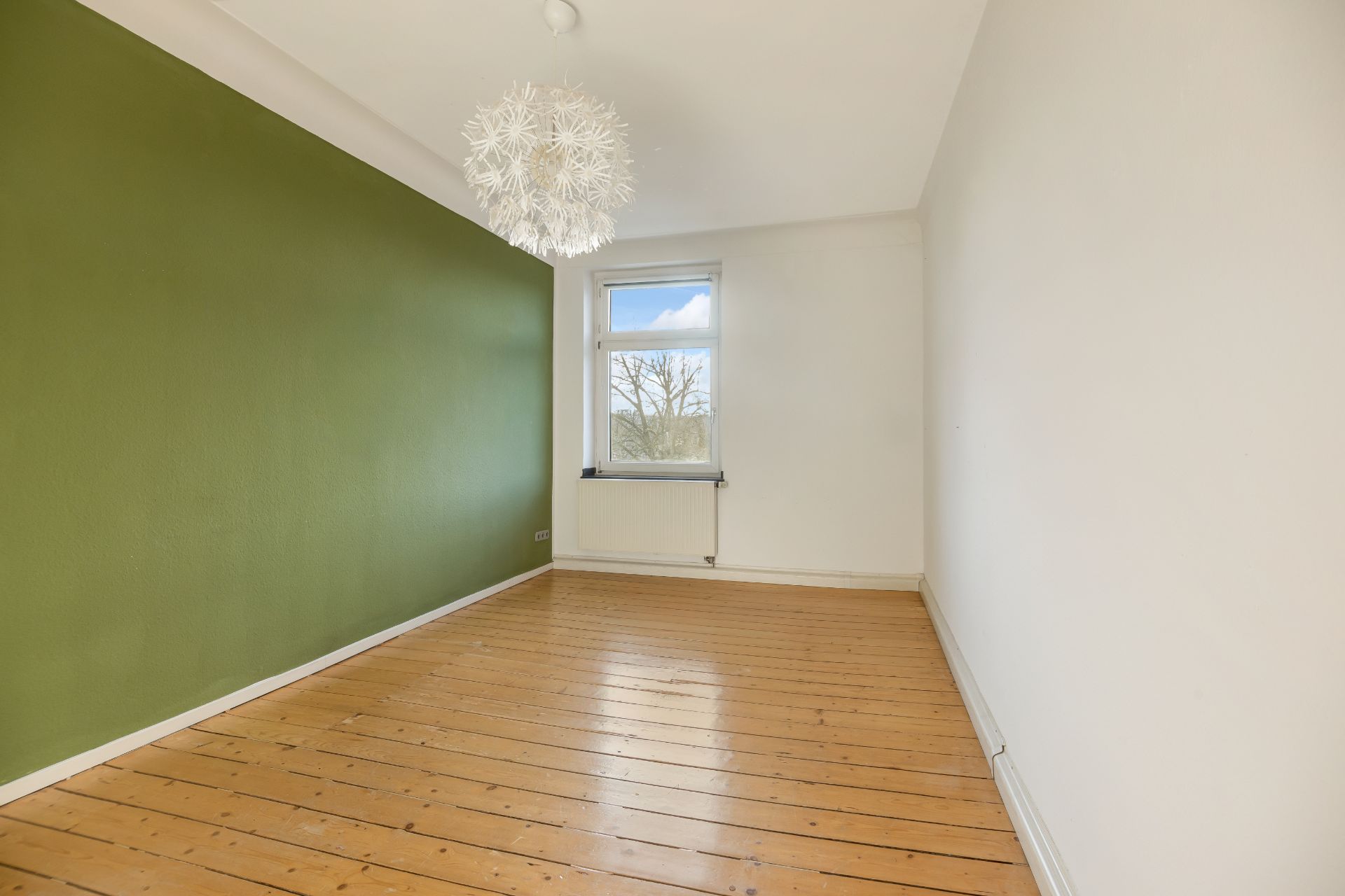 Leerer Raum mit Holzboden, grüner Wand und modernem Kronleuchter, Fenster mit Blick nach draußen.