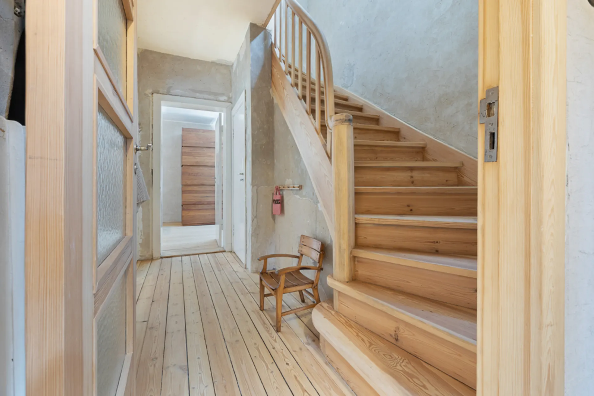 Flur eines Hauses mit Holztreppe und kleinem Holzstuhl, unbehandelte Wände.