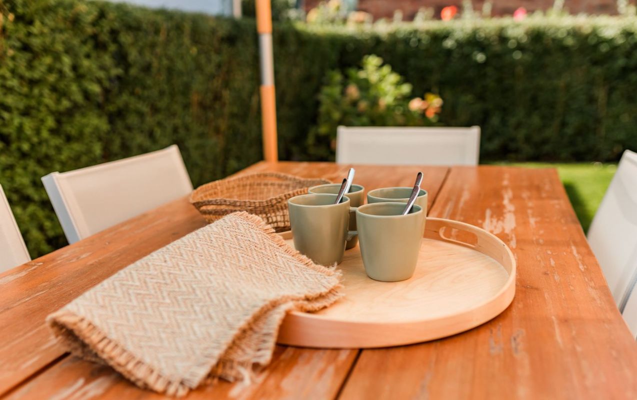 Holztisch im Garten mit Tablett, vier Tassen, Löffeln und beiger Decke.