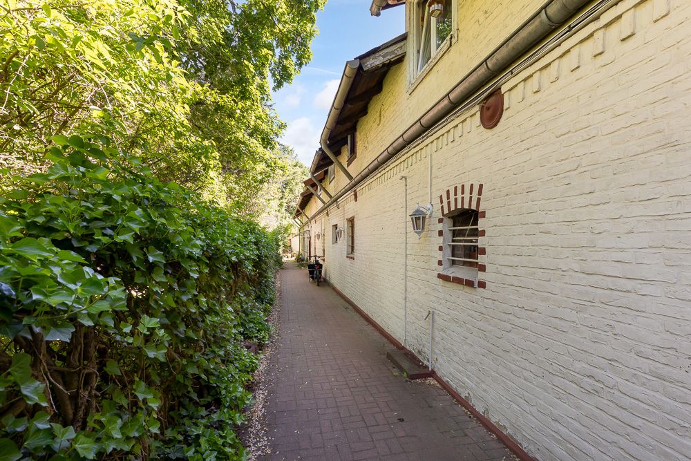 Gepflasterter Weg neben hellgelbem Haus mit Fenstern, Hecke links, Bäume im Hintergrund.