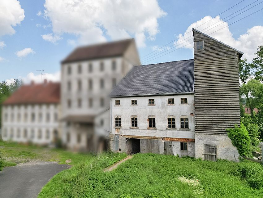 Altes zweistöckiges Gebäude mit Holzanbau und mehreren Fenstern, umgeben von Grünfläche.