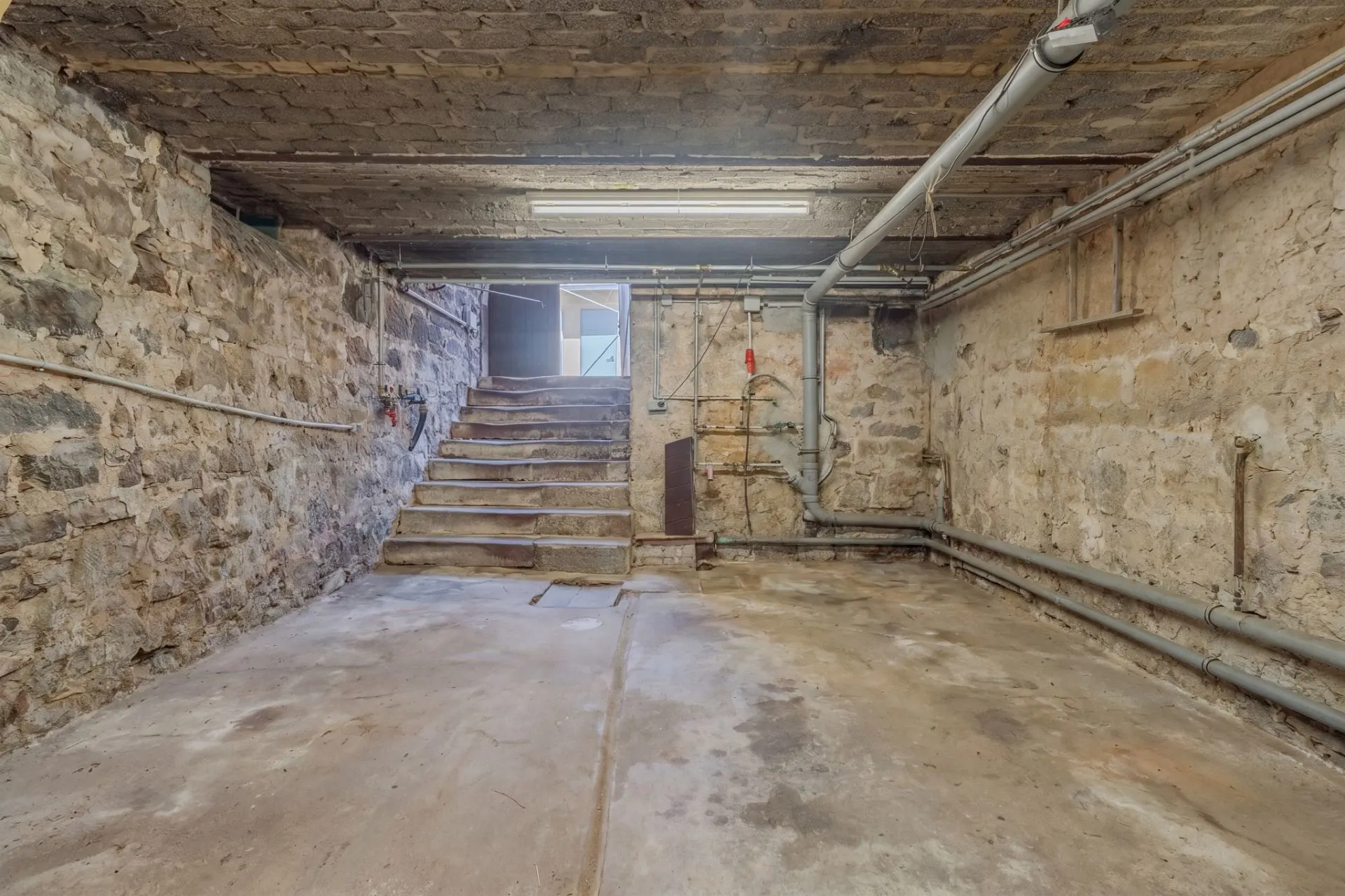 Kellerraum mit Steinwänden, Betonboden und sichtbaren Rohren, Treppe führt nach oben.