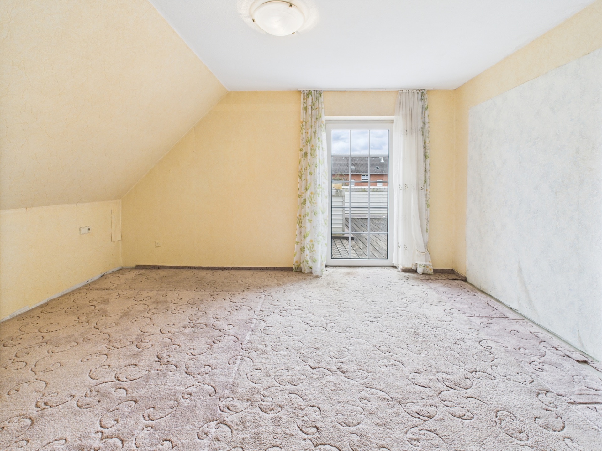Leeres Zimmer mit gemustertem Teppich und Fenstertür zu einem Balkon.