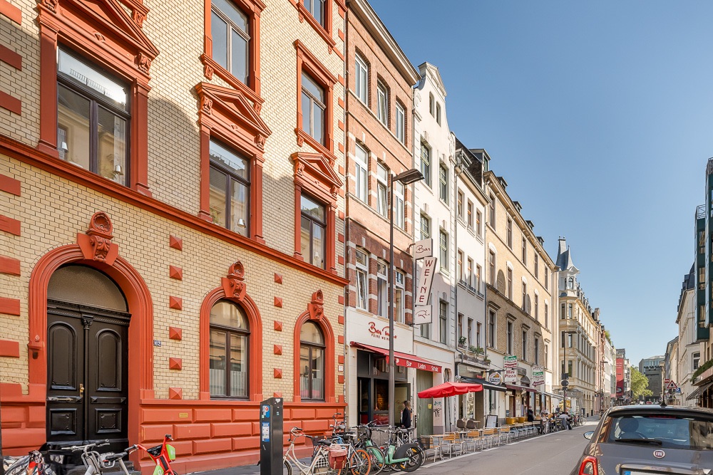 Mehrstöckiges Wohnhaus mit gelber und roter Fassade an einer Straße mit geparkten Fahrrädern und Autos.