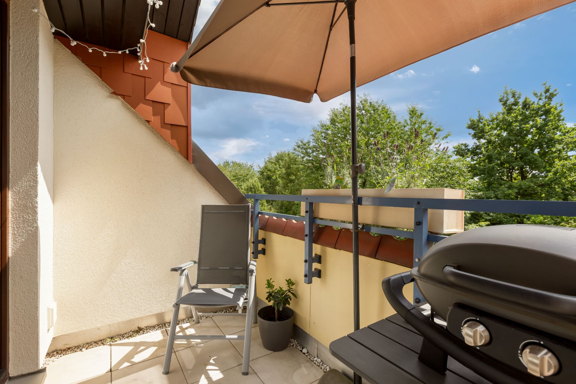 Kleiner Balkon mit Stuhl, Grill, Sonnenschirm und Blumentopf, umgeben von Bäumen.