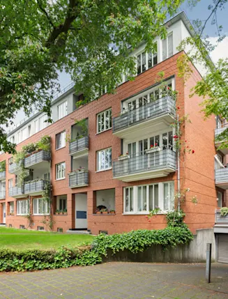 Mehrstöckiges Wohnhaus aus Backstein mit Balkonen, von Bäumen und viel Grün umgeben.