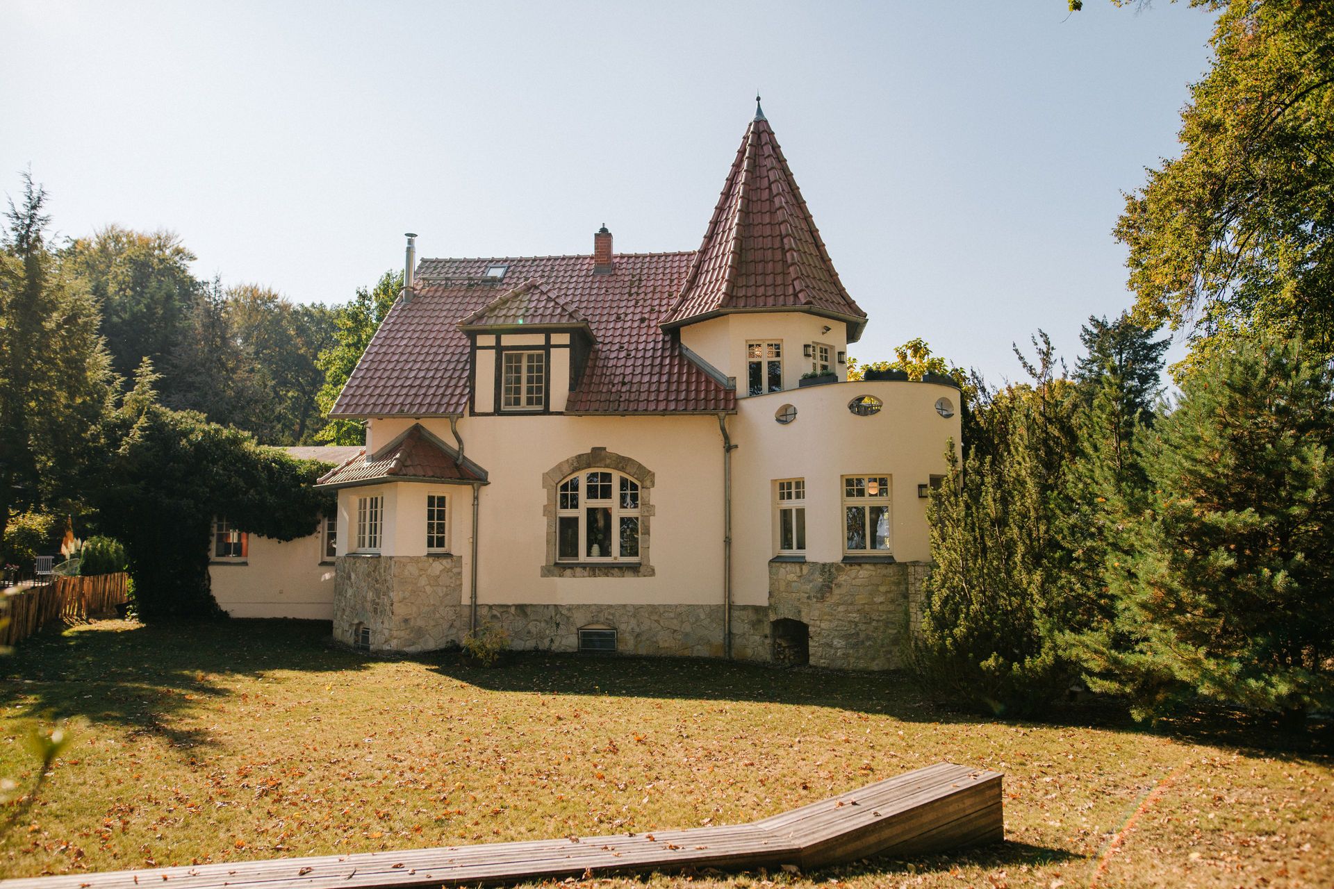 Großes Wohnhaus mit rotem Dach und Turm, umgeben von Garten und Bäumen.