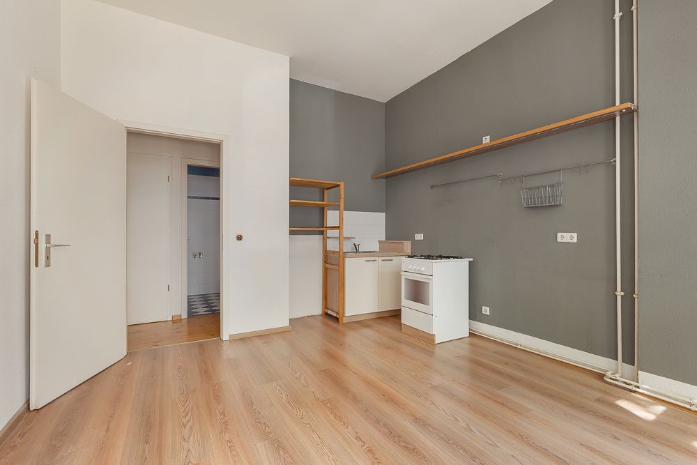 Heller Raum mit Holzboden, kleiner Küchenzeile, Regal und Gasherd an einer grauen Wand.