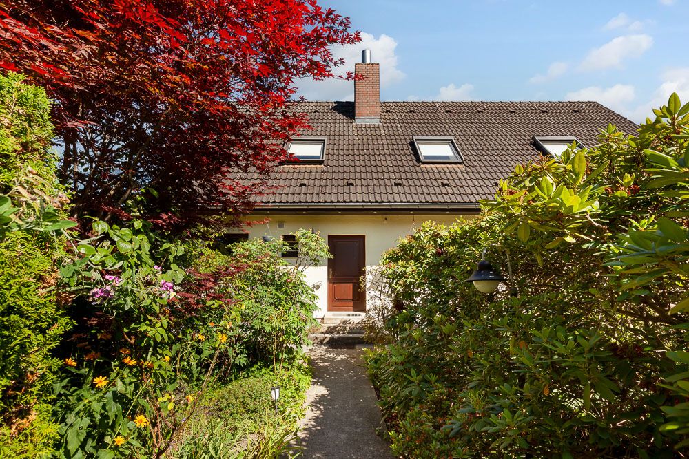 Einfamilienhaus mit braunem Dach, Weg zum Eingang, umgeben von Bäumen und Sträuchern.