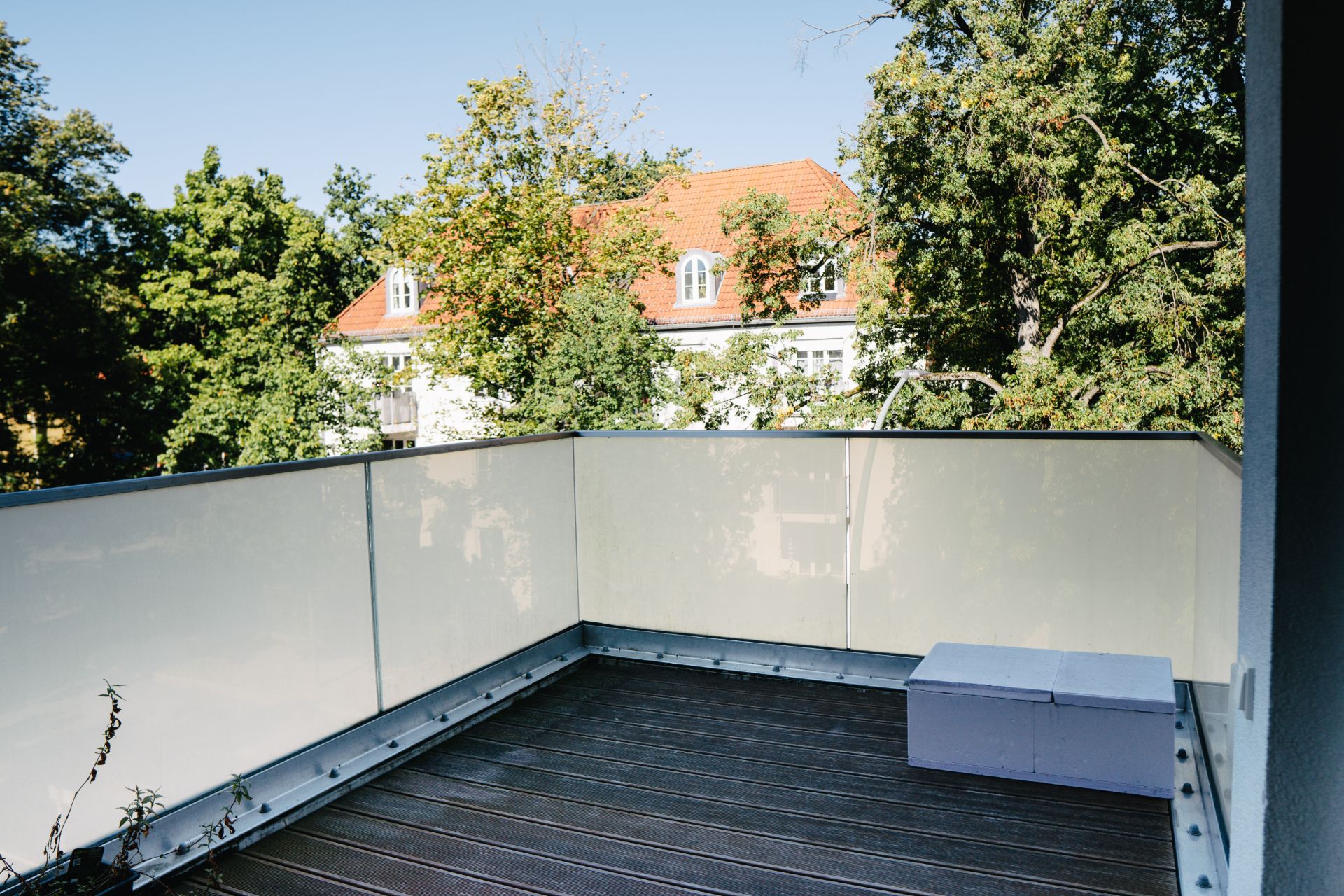 Balkon mit Holzboden, einem kleinen Kasten und Blick auf Bäume und ein Haus mit rotem Dach.