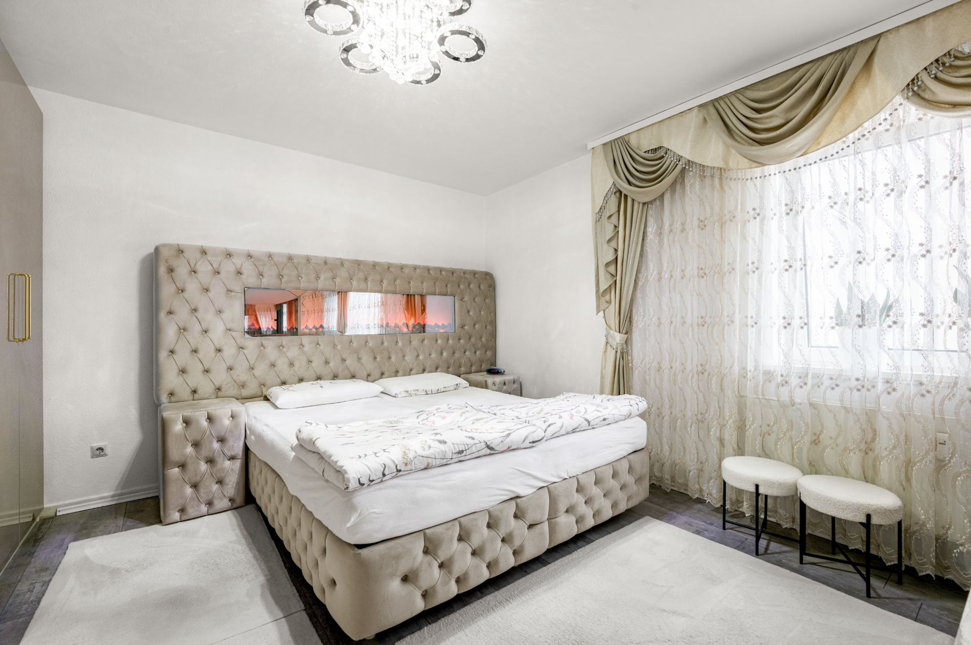 Helles Schlafzimmer mit großem gepolsterten Bett, Kronleuchter und großen Fenstern mit Vorhängen.