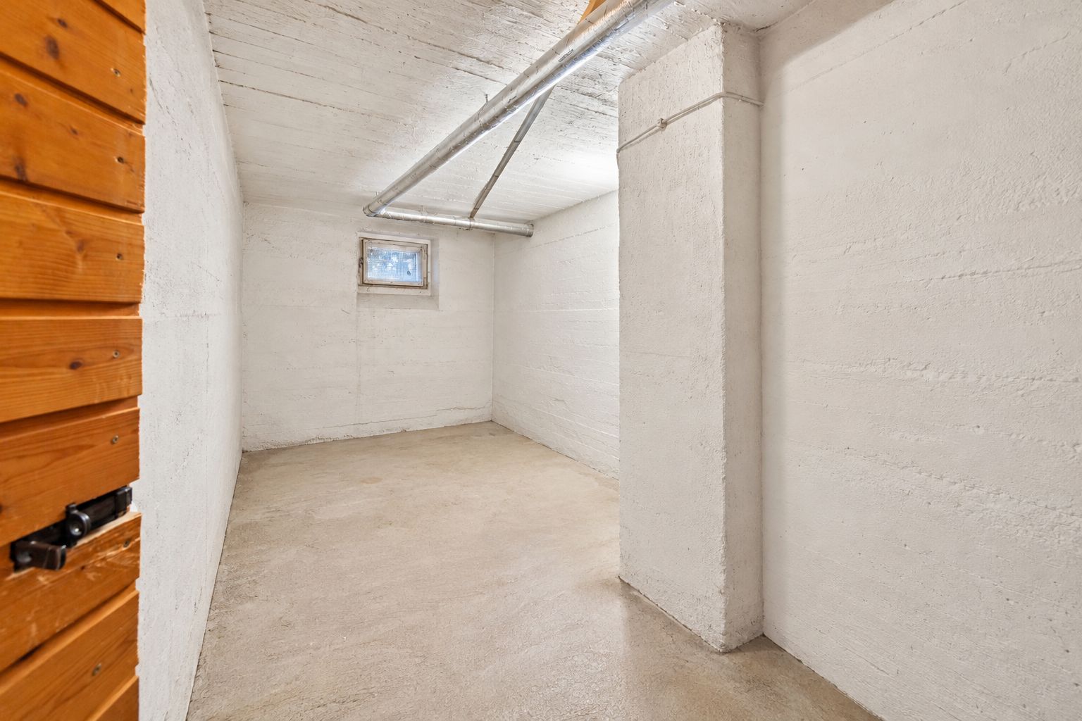 Kleiner, leerer Kellerraum mit weißen Wänden, einem Fenster und sichtbaren Rohren an der Decke.