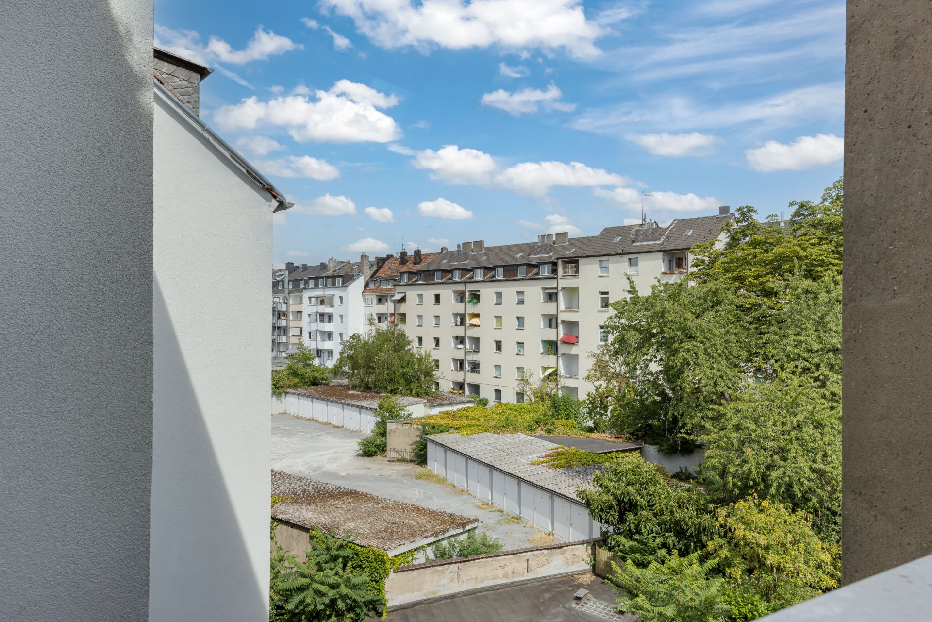 Blick von einem Balkon auf Wohngebäude, Bäume und Garagen bei blauem Himmel.