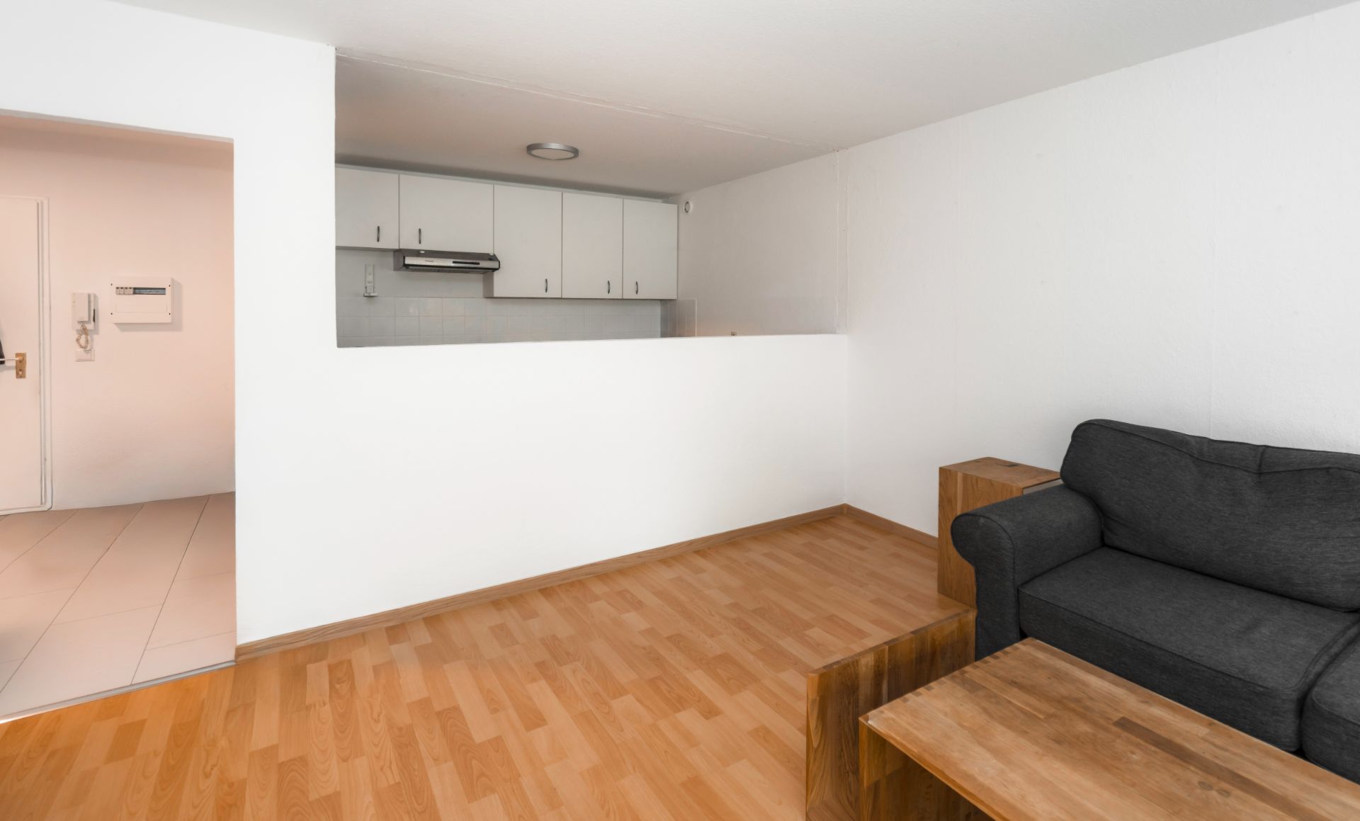 Wohnzimmer mit Holzfußboden, grauem Sofa, Holztisch und offener Küche im Hintergrund.