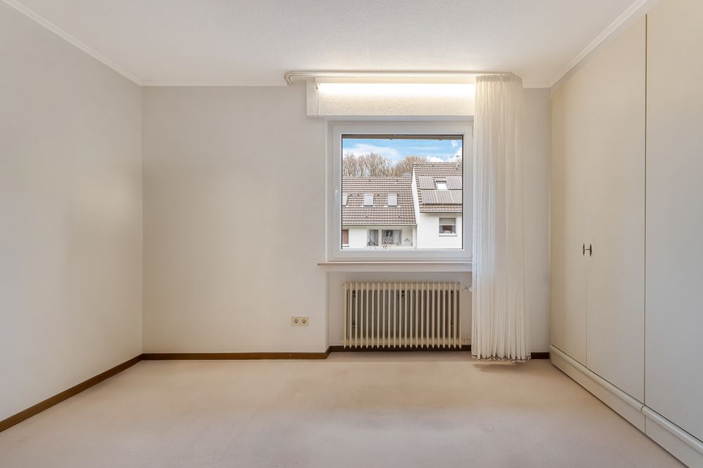 Leeres Zimmer mit Teppichboden, großem Fenster, Heizung und Einbauschrank.