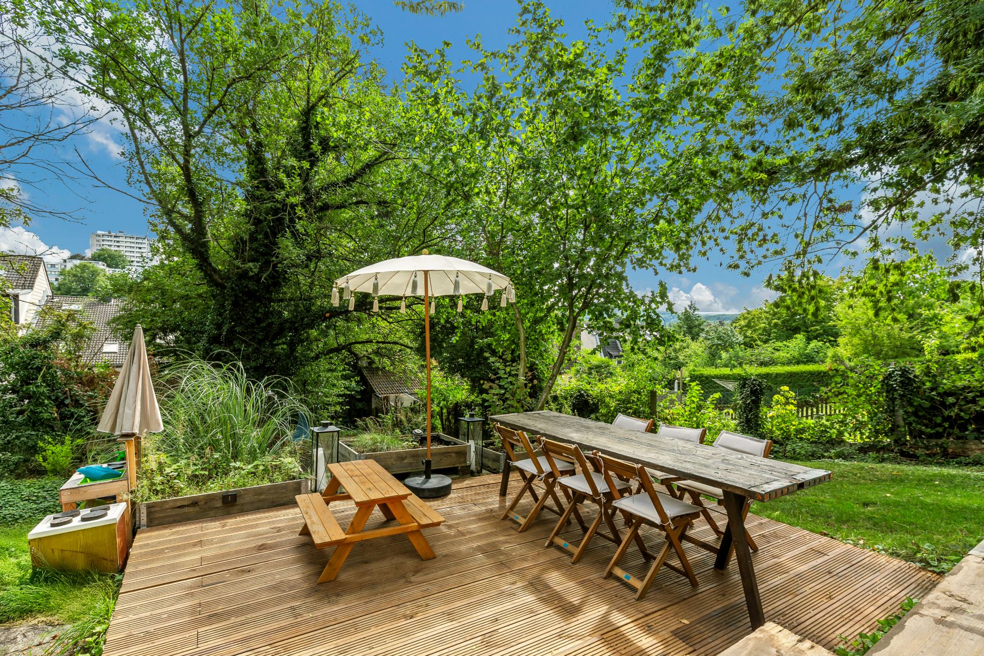 Holzterrasse mit Tisch, Stühlen, Bank und Sonnenschirm, umgeben von Bäumen und Grün.
