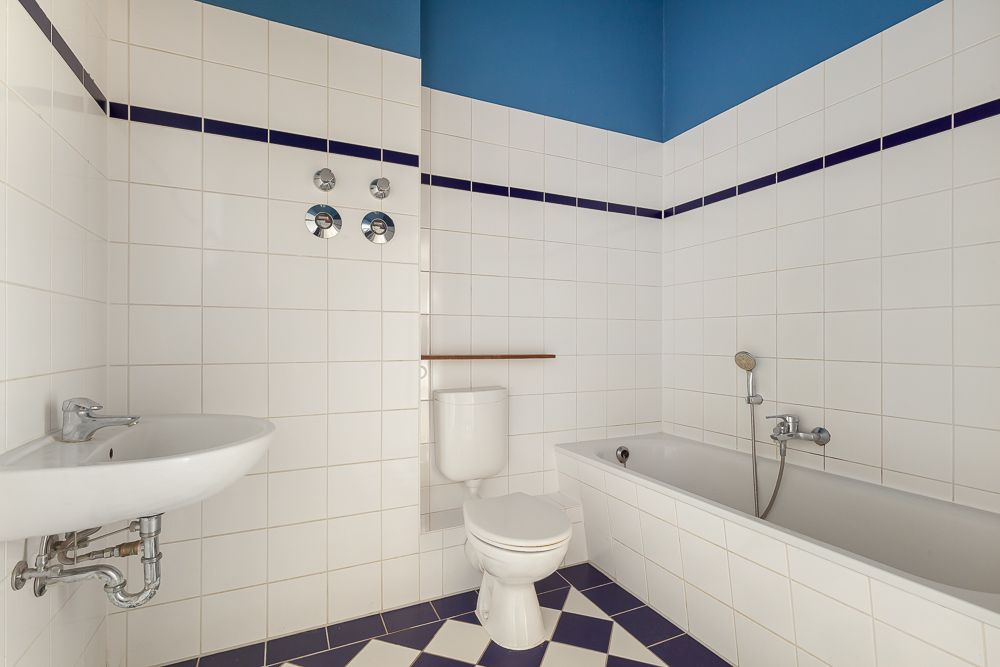 Weiß gefliestes Badezimmer mit Badewanne, Toilette und Waschbecken. Blaue Wand und Bodenfliesen.