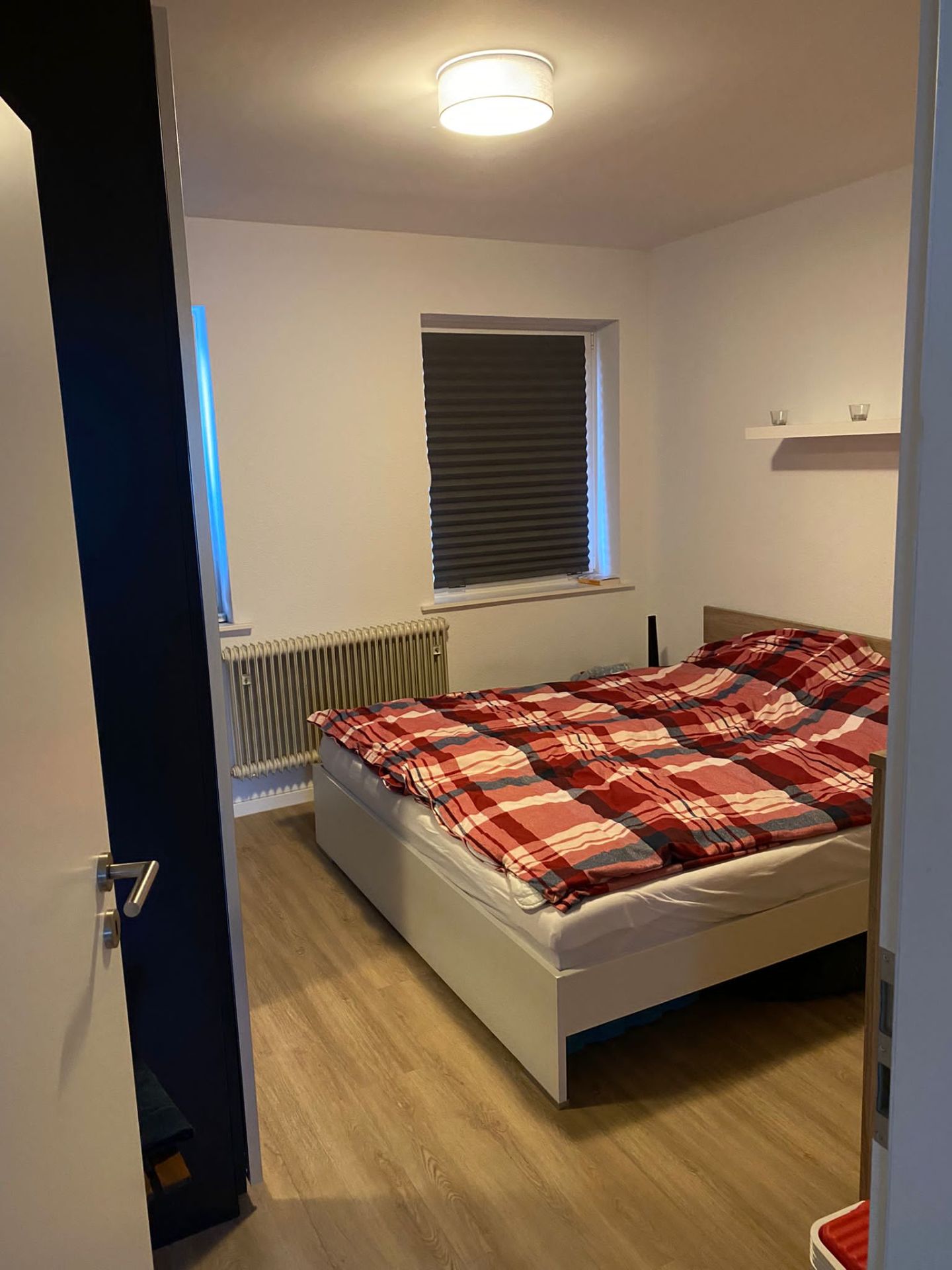 Schlafzimmer mit Doppelbett, kariertem Bettbezug, Fenster mit Rollladen und Parkettboden.