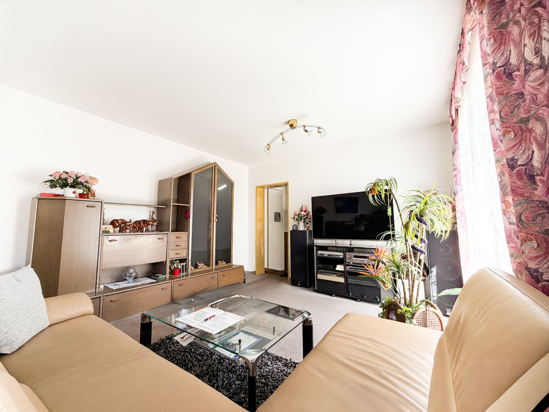 Helles Wohnzimmer mit beiger Eckcouch, Glastisch, TV, Pflanzen und Schrankwand.