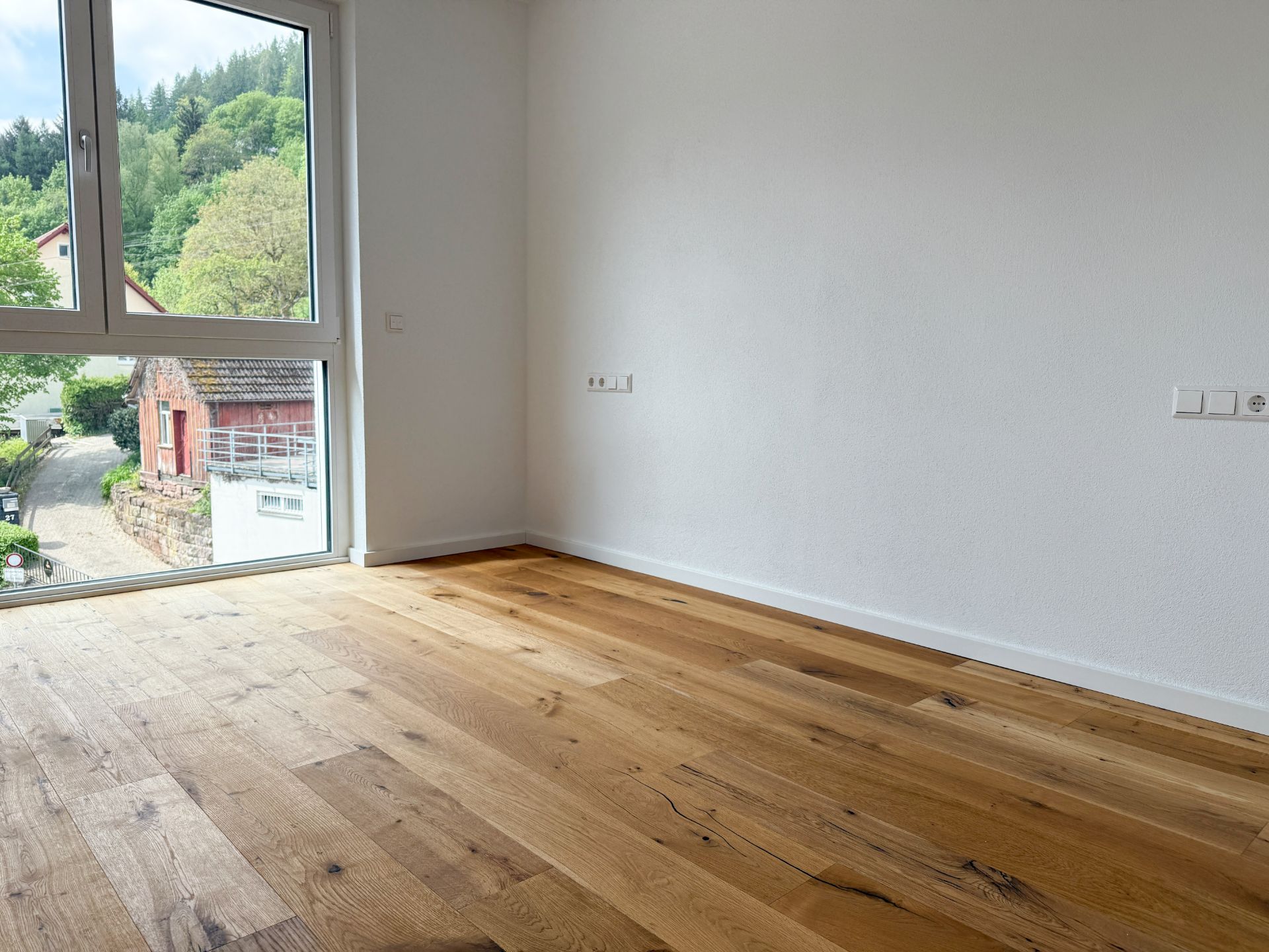 Leerer Raum mit Holzfußboden und großem Fenster, Blick auf grüne Landschaft außerhalb.