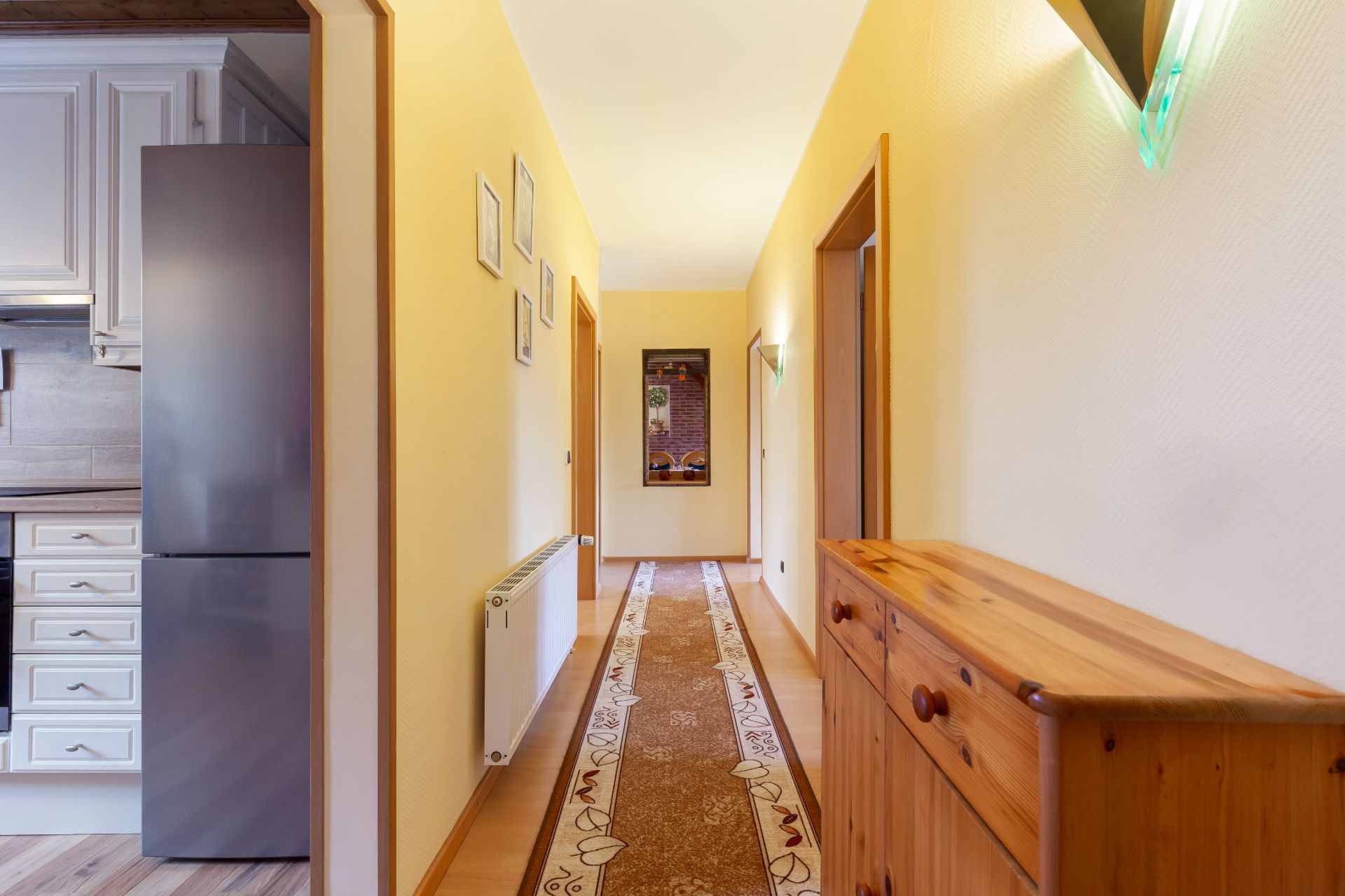 Heller Flur mit Teppichläufer, Kommode und Blick in eine Küche mit Kühlschrank.