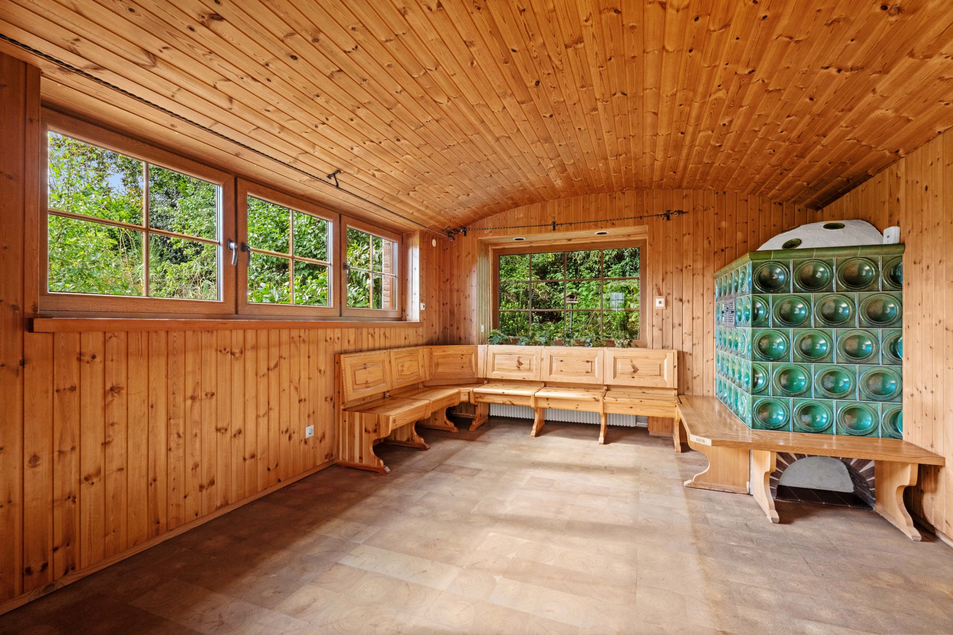 Holzvertäfelter Raum mit Eckbank, großen Fenstern und grünem Kachelofen.