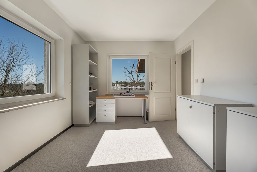 Helles Zimmer mit Schreibtisch, Regal, Fenster, Tür und weißen Schränken.